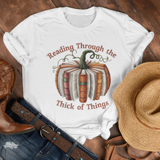 White t-shirt with pumpkin design and text, paired with cowboy boots, denim shorts, a straw hat, and brown purse on wooden surface. Text is Reading Through the Thick of Things.